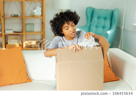 African girl unpacking delivery looking in box. Happy woman opening carton box. Female getting parcel looking at delivered goods items. Satisfied client positive feedback quick express deliver African girl unpacking delivery looking in box. Happy woman opening carton box. Female getting parcel looking at delivered goods items. Satisfied client positive feedback quick express deliver 106455634