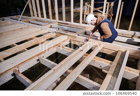 Man worker building wooden frame house on pile foundation. Carpenter hammering nail into wooden joist, using hammer. Carpentry concept. 106469540