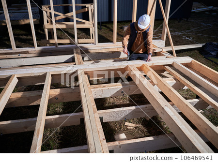 Man worker building wooden frame house on pile foundation. Carpenter hammering nail into wooden plank, using hammer. Carpentry concept. Man worker building wooden frame house on pile foundation. Carpenter hammering nail into wooden plank, using hammer. Carpentry concept. 106469541