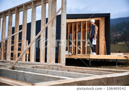 Male designer building wooden frame house. Man builder standing on construction site in safety helmet, with construction documentation inspecting quality of work. 106469542