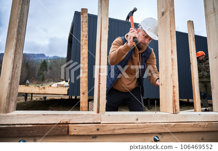 Man worker building wooden frame house. Carpenter hammering nail into wooden joist, using hammer. Carpentry concept. Man worker building wooden frame house. Carpenter hammering nail into wooden joist, using hammer. Carpentry concept. 106469552