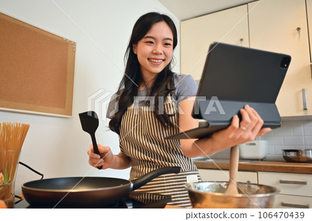 Pretty young woman wearing apron cooking in the kitchen and following an online recipe on digital tablet Pretty young woman wearing apron cooking in the kitchen and following an online recipe on digital tablet 106470439