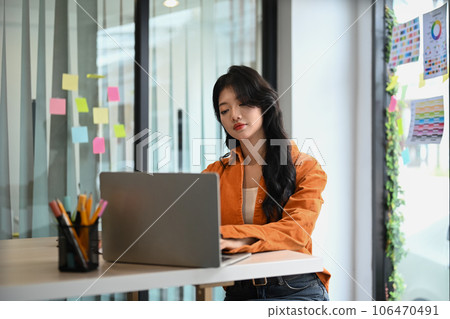 Young creative woman in orange shirt sitting in bright office and working on design project on laptop computer 106470491