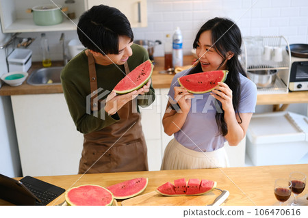 Moment of loving young asian couple wearing aprons standing behind wooden kitchen tabletop and eating sweet watermelon Moment of loving young asian couple wearing aprons standing behind wooden kitchen tabletop and eating sweet watermelon 106470616
