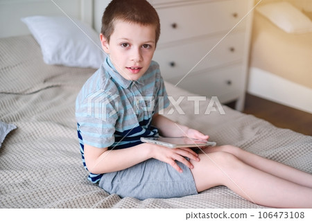 Caucasian school-aged boy in a shirt and shorts, seated on a couch with a tablet in hand, smiling Caucasian school-aged boy in a shirt and shorts, seated on a couch with a tablet in hand, smiling 106473108