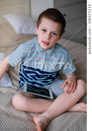 Caucasian school-aged boy in a shirt and shorts, seated on a couch with a tablet in hand, smiling 106473114
