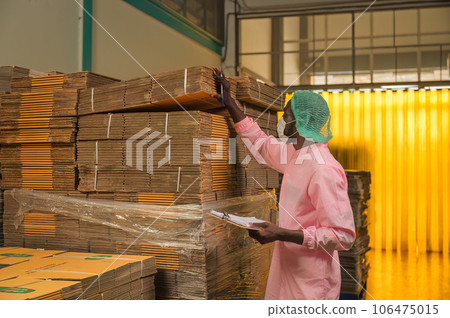 African male employee checking with clipboard at stacked cardboard package stuck in shipping warehouse at manufacturing factory African male employee checking with clipboard at stacked cardboard package stuck in shipping warehouse at manufacturing factory 106475015