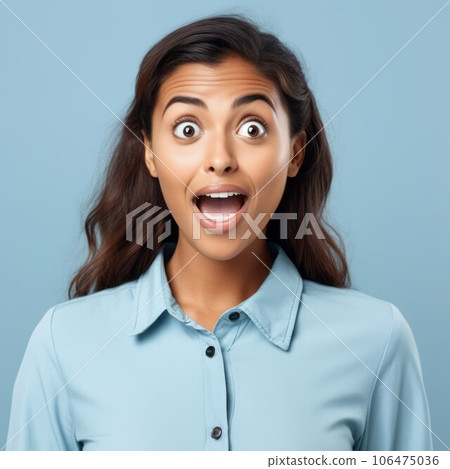 Portrait of a surprised Latin American woman with big eyes and an open mouth. Closeup face of an amazed adult Latin girl on a blue background. Astonished grownup in a blue shirt looking at the camera. 106475036