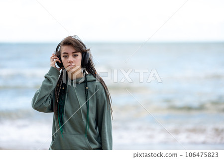 Close-up of teenager girl with dreadlocks listening to music with headphones on the seashore during the daytime. 106475823