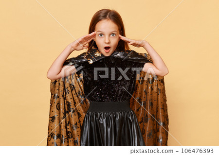 Shocked surprised little girl wearing witch costume isolated over beige background looking at camera with big eyes and open mouth amazing halloween party 106476393