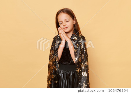 Sleepy tired little girl wearing witch costume isolated over beige background standing leaning on her hands keeps eye closed halloween party. 106476394