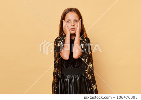 Shocked scared little girl wearing witch costume isolated over beige background keeps hands on cheeks looking with big eyes at something scary halloween celebration. 106476395