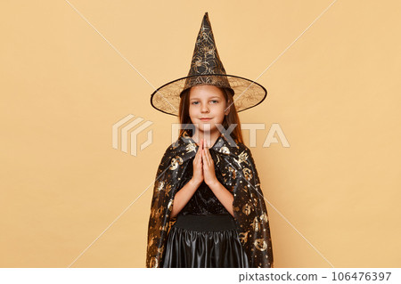 Cute smiling little girl wearing witch costume and carnival cone hat isolated over beige background standing with hands together praying her magic spell worked. Cute smiling little girl wearing witch costume and carnival cone hat isolated over beige background standing with hands together praying her magic spell worked. 106476397