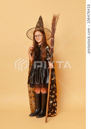 Full length portrait of little girl with long hair black cloak clothing with broom isolated over beige background looking at camera celebrating halloween at school party. Full length portrait of little girl with long hair black cloak clothing with broom isolated over beige background looking at camera celebrating halloween at school party. 106476416