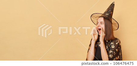 Caucasian young woman wearing witch costume and carnival cone hat celebrating halloween isolated over beige background screaming loud making announcement looking at advertisement area. Caucasian young woman wearing witch costume and carnival cone hat celebrating halloween isolated over beige background screaming loud making announcement looking at advertisement area. 106476475
