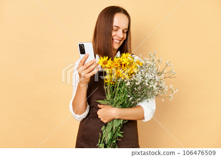 Cheerful young caucasian woman florist wearing brown apron holding flowers isolated over beige background using mobile phone looking at bouquet with smile 106476509