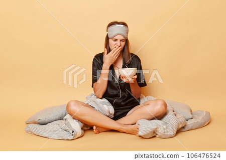 Displeased sad young girl sitting floor with blanket wearing black pajama and sleep eye mask holding plate in hands isolated over beige background felling nausea bed smell of breakfast meal 106476524