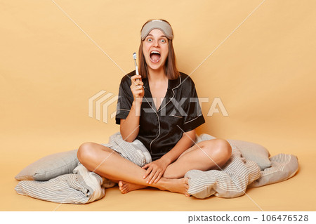 Excited overjoyed young girl sitting floor with blanket wearing black pajama and sleep eye mask isolated over beige background screaming with amazement holding toothbrush. Excited overjoyed young girl sitting floor with blanket wearing black pajama and sleep eye mask isolated over beige background screaming with amazement holding toothbrush. 106476528