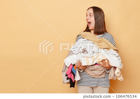 Excited brunette woman holding pile of clothes in hands isolated over beige background looking away at copy space for promotion advertisement area screaming with happiness. Excited brunette woman holding pile of clothes in hands isolated over beige background looking away at copy space for promotion advertisement area screaming with happiness. 106476534