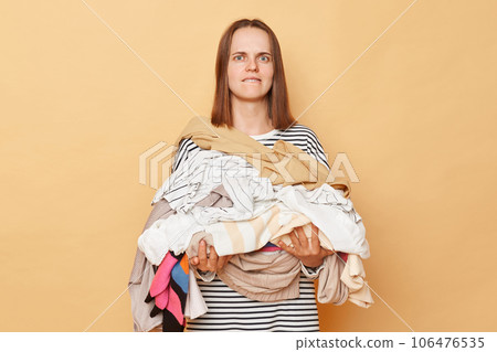 Confused brunette woman holding pile of clothes in hands isolated over beige background looking at camera with confused expression biting her lip. Confused brunette woman holding pile of clothes in hands isolated over beige background looking at camera with confused expression biting her lip. 106476535