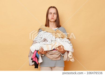 Tired exhausted brunette woman holding pile of clothes in hands isolated over beige background looking at camera with crossed eyes blowing cheeks doing lots house work. 106476536