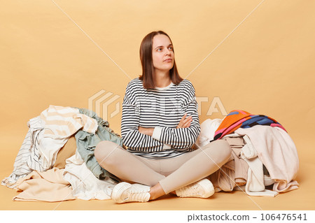 Pensive Caucasian woman posing near heap of multicolored unsorted clothes isolated over beige background sorting her wardrobe thinking where to put unnecessary clothing 106476541