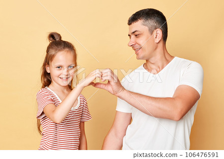 Cute charming positive man and little kid girl standing together isolated over beige background showing heart shape with hand expressing love and positive emotions. Cute charming positive man and little kid girl standing together isolated over beige background showing heart shape with hand expressing love and positive emotions. 106476591