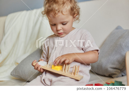 Child's educational playtime. Playful games for curious minds. Creative learning environment. Focused wavy haired blonde toddler baby playing wooden educational toys at home interior. Child's educational playtime. Playful games for curious minds. Creative learning environment. Focused wavy haired blonde toddler baby playing wooden educational toys at home interior. 106476779