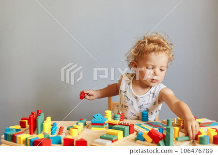 Childhood curiosity and discovery. Playful games for curious minds. Cute creative wavy haired blonde toddler baby playing wooden educational toys isolated over gray background Childhood curiosity and discovery. Playful games for curious minds. Cute creative wavy haired blonde toddler baby playing wooden educational toys isolated over gray background 106476789