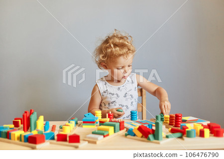 Exploring educational activities. Playful games in daycare. Kindergarten room of wonder. Focused wavy haired blonde infant baby playing wooden educational toys isolated over gray background Exploring educational activities. Playful games in daycare. Kindergarten room of wonder. Focused wavy haired blonde infant baby playing wooden educational toys isolated over gray background 106476790