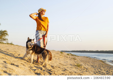 Woman in hat and sunglasses with her dog enjoying sunset on the beach 106481590