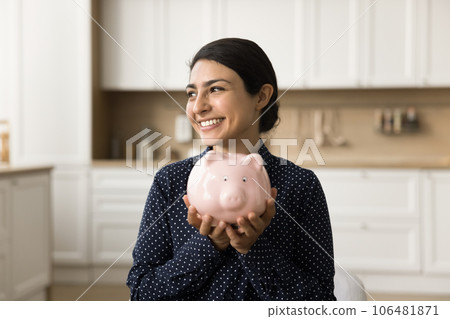 Cheerful young Indian woman holding toy ceramic pig with cash 106481871