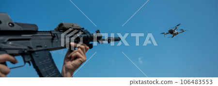 A man aims to shoot a rifle at a flying drone against a blue sky. A man aims to shoot a rifle at a flying drone against a blue sky. 106483553