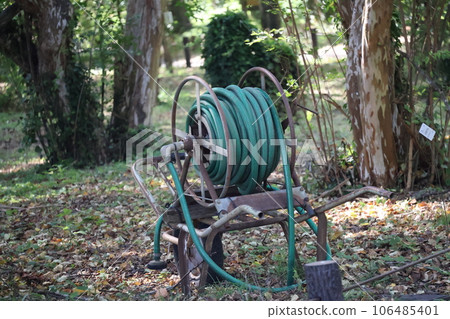 Scenery of a cart with a hose spraying water in a park with many plants Scenery of a cart with a hose spraying water in a park with many plants 106485401