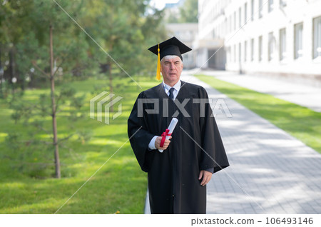 Serious old man in graduation gown holding diploma outdoors. 106493146