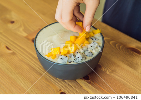 Woman cooks smoothie bowls made with mango, banana, granola, grated coconut, dragon fruit, chia seeds and mint on wooden background. Concept fruits, vitamins Woman cooks smoothie bowls made with mango, banana, granola, grated coconut, dragon fruit, chia seeds and mint on wooden background. Concept fruits, vitamins 106496841