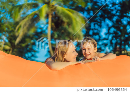 Summer lifestyle portrait of mother and son sitting on the orange inflatable sofa on the beach of tropical island. Relaxing and enjoying life on air bed Summer lifestyle portrait of mother and son sitting on the orange inflatable sofa on the beach of tropical island. Relaxing and enjoying life on air bed 106496864