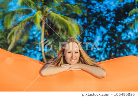 Summer lifestyle portrait of pretty girl sitting on the orange inflatable sofa on the beach of tropical island. Relaxing and enjoying life on air bed 106496886
