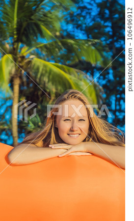 Summer lifestyle portrait of pretty girl sitting on the orange inflatable sofa on the beach of tropical island. Relaxing and enjoying life on air bed VERTICAL FORMAT for Instagram mobile story or 106496912