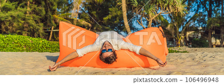 Summer lifestyle portrait of man sitting on the orange inflatable sofa on the beach of tropical island. Relaxing and enjoying life on air bed BANNER, LONG FORMAT Summer lifestyle portrait of man sitting on the orange inflatable sofa on the beach of tropical island. Relaxing and enjoying life on air bed BANNER, LONG FORMAT 106496948