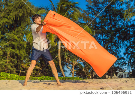 Summer lifestyle portrait of man inflates an inflatable orange sofa on the beach of tropical island. Relaxing and enjoying life on air bed Summer lifestyle portrait of man inflates an inflatable orange sofa on the beach of tropical island. Relaxing and enjoying life on air bed 106496951