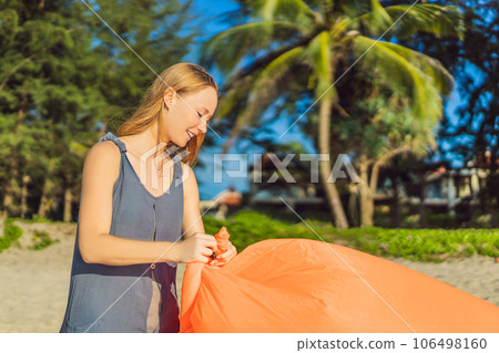 Summer lifestyle portrait of woman inflates an inflatable orange sofa on the beach of tropical island. Relaxing and enjoying life on air bed Summer lifestyle portrait of woman inflates an inflatable orange sofa on the beach of tropical island. Relaxing and enjoying life on air bed 106498160