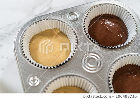 Cupcake foil liners are being meticulously filled with both chocolate and vanilla batter, setting the stage for the baking of deliciously diverse birthday cupcakes. Cupcake foil liners are being meticulously filled with both chocolate and vanilla batter, setting the stage for the baking of deliciously diverse birthday cupcakes. 106498730