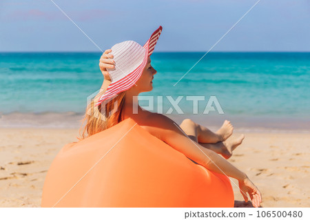 Summer lifestyle portrait of pretty girl sitting on the orange inflatable sofa on the beach of tropical island. Relaxing and enjoying life on air bed 106500480