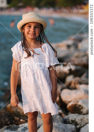 Portrait of adorable little girl in white dress and knitted hat stand on rocks by the sea. 106503372