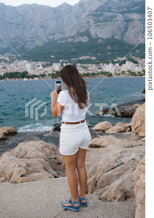 Back view of a woman taking a video on her phone of a sea sunset. Landscape of mountains Back view of a woman taking a video on her phone of a sea sunset. Landscape of mountains 106503407