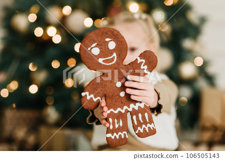 Merry Christmas and Happy Holidays. Child girl eating big iced ingerBread man cookies under Christmas tree Waiting for Christmas. Celebration New Year. 106505143