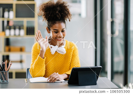 Attractive young African student studying at the college library, sitting at the desk, using a laptop computer, tablet and headphones having a video chat. Attractive young African student studying at the college library, sitting at the desk, using a laptop computer, tablet and headphones having a video chat. 106505300