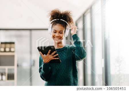Attractive young African student studying at the college library, sitting at the desk, using a laptop computer, tablet and headphones having a video chat. Attractive young African student studying at the college library, sitting at the desk, using a laptop computer, tablet and headphones having a video chat. 106505451