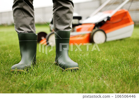 Cropped photo of male gardener legs in rubber boots stands on cut green grass lawn at backyard of house. Modern electric cordless lawnmower on back. Landscaping industry theme. 106506446
